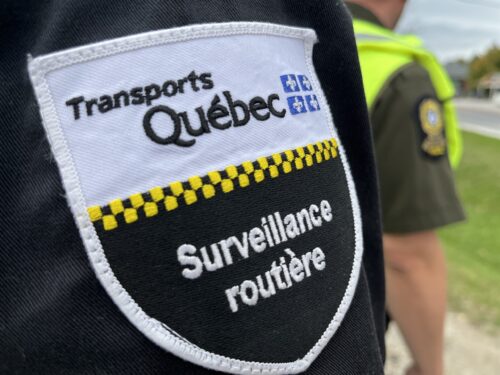 Le logo de Transports Québec sur le bras d'un agent chargé de la surveillance routière au Québec, avec un policier de la Sûreté du Québec en arrière-plan.