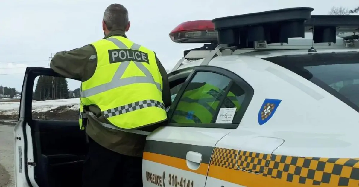 Un policier de la Sûreté du Québec vu de dos portant un dossard de sécurité jaune se tient debout derrière la porte de son véhicule de patrouille en bordure de route.