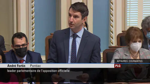 Le député du Parti libéral du Québec, André Fortin, s’exprime debout à l’Assemblée nationale du Québec, tenant des documents lors d’une période de débats parlementaires. D’autres élus sont visibles assis derrière lui, dans un cadre institutionnel formel.