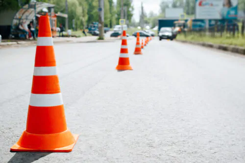 Traffic safety cones on asphalt of road. Selective focus on striped orange-white warning signs, cars bokeh