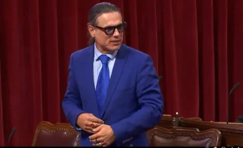 Un homme autochtone aux cheveux noirs se tient debout dans l'enceinte du Sénat pour parler. Il pointe la tête vers sa gauche pour parler à quelqu'un. Il porte une chemise blanche, un cravate et un complet bleus, ainsi que des bagues aux doigts.