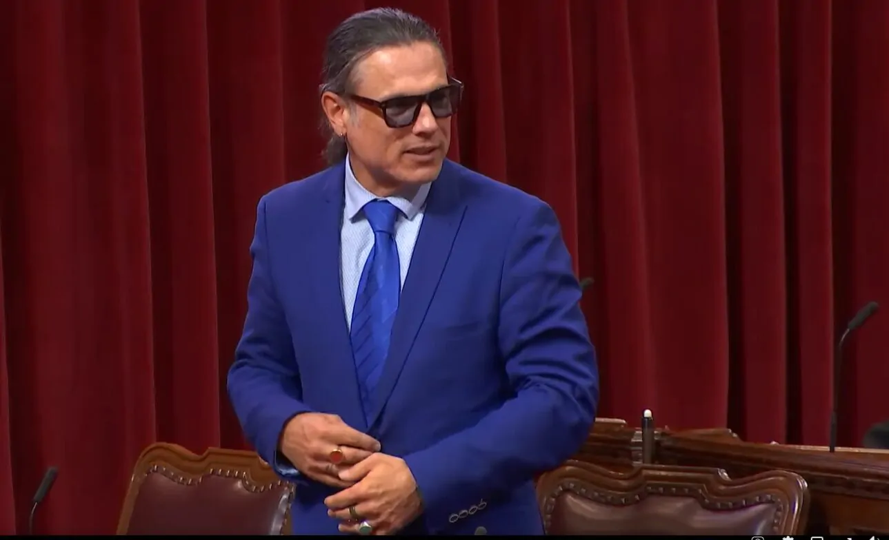Un homme autochtone aux cheveux noirs se tient debout dans l'enceinte du Sénat pour parler. Il pointe la tête vers sa gauche pour parler à quelqu'un. Il porte une chemise blanche, un cravate et un complet bleus, ainsi que des bagues aux doigts.