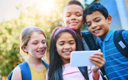 Un groupe d'élèves portant des sacs à dos, composé de deux petites filles et de deux petits garçons, regarde l'écran d'un téléphone intelligent en souriant. Derrière eux, il y a des arbres.