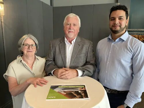 Une femme et deux hommes se tiennent debout derrière une table avec une nappe blanche. Un document est posé sur la table.