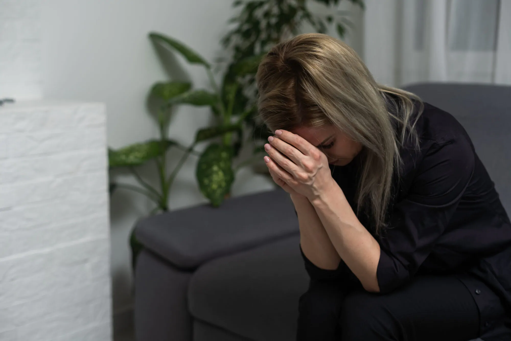 Une femme triste habillée en noir et assise sur un sofa tient sa tête avec ses mains jointes en signe de prière. En arrière-plan, on voit un fond blanc avec une plante.
