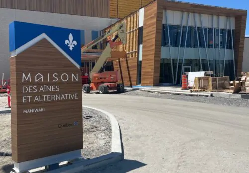 Une pancarte en bois avec le symbole du gouvernement du Québec, un lys blanc, indique la Maison des aînés et alternative de Maniwaki. On voit le bâtiment en construction à l'arrière-plan. Il est en voie d'être terminé.