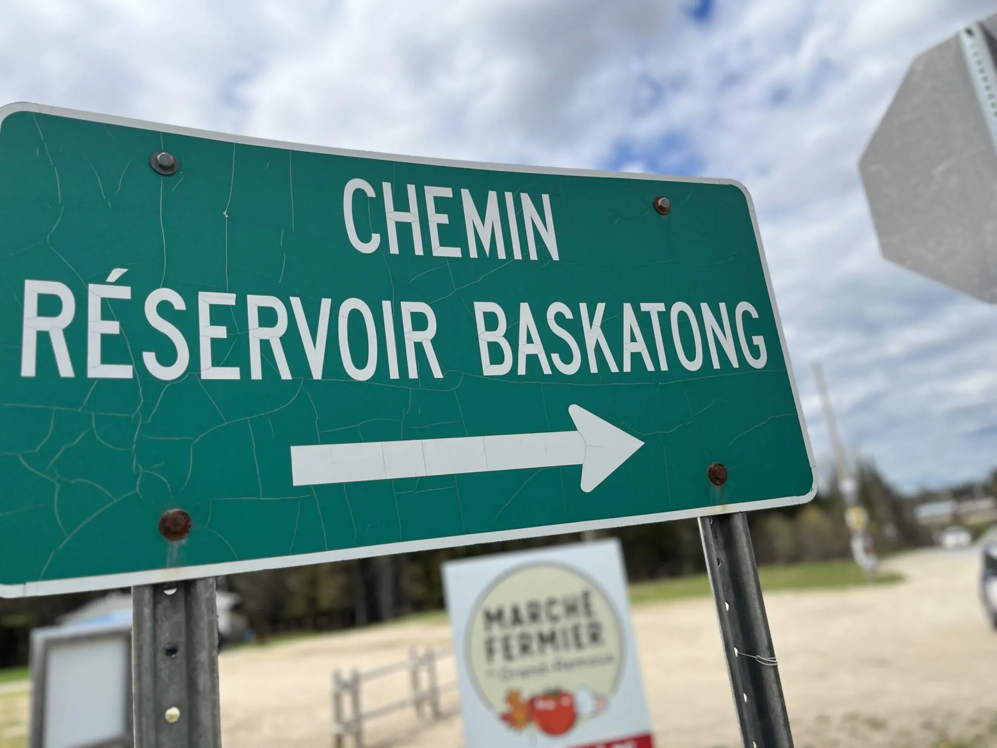 Une pancarte de signalisation verte avec une flèche blanche qui indique la direction à suivre aux automobilistes. Le panneau indique le chemin du réservoir Baskatong. On apperçoit derrière un stationnement de terre, des arbres et un ciel variable.