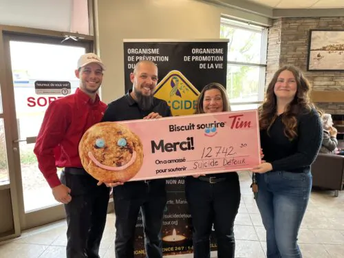 Deux hommes et deux femmes tiennent dans leurs mains une banderole de la campagne des biscuits sourire. Le montant indiqué par le chèque est de 12 742$. Derrière eux, il y a une bannière de Suicide Détour. Le groupe se trouve à l'intérieur du restaurant Tim Hortons de Maniwaki.