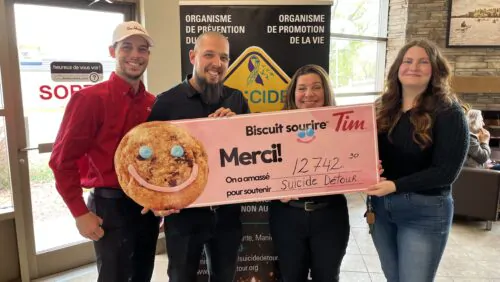 Deux hommes et deux femmes tiennent dans leurs mains une banderole de la campagne des biscuits sourire. Le montant indiqué par le chèque est de 12 742$. Derrière eux, il y a une bannière de Suicide Détour. Le groupe se trouve à l'intérieur du restaurant Tim Hortons de Maniwaki.
