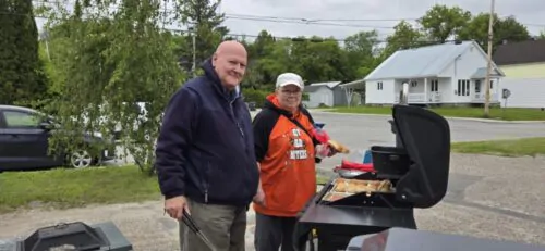 Un homme avec polar bleu et une femme avec polar orangé et casquette blanche, devant un BBQ faisant cuire pains hamburger