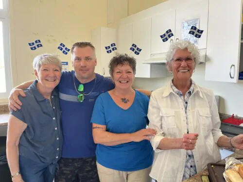 Un groupe de trois femmes et un homme posent pour une photo de groupe dans une cuisine communautaire blanche, avec des petits drapeaux du Québec sur la tête.