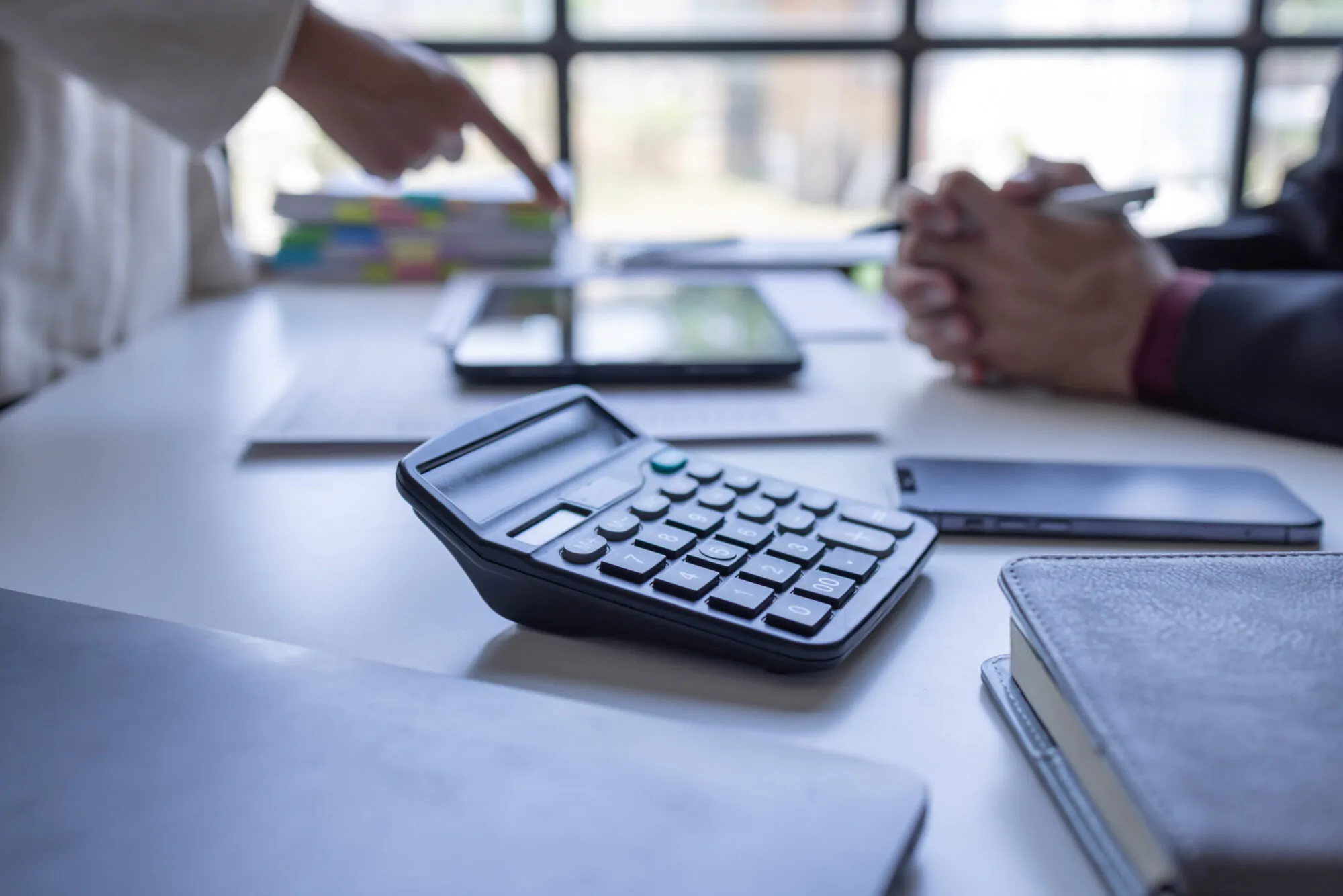 Une calculatrice et une pile de documents sont posés sur un bureau alors que deux personnes discutent en arrière-plan.