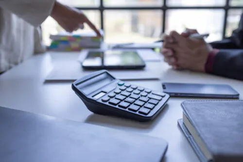 Une calculatrice et une pile de documents sont posés sur un bureau alors que deux personnes discutent en arrière-plan.