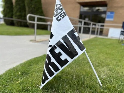 Un petit drapeau blanc avec les inscriptions " en grève " et " CSN " dessus est planté dans de la pelouse verte près d'une allée menant à un bâtiment en brique.