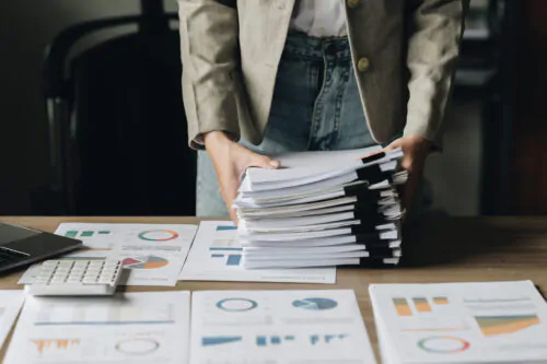 Une femme tient dans ses mains une pile de dossiers posés sur un bureau. Sur le bureau, il y a des documents avec des graphiques.