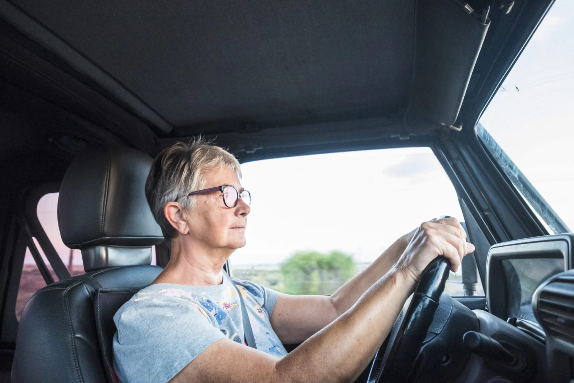 Une femme avec les cheveux gris et portant des lunettes et un tee-shirt clair conduit une voiture.
