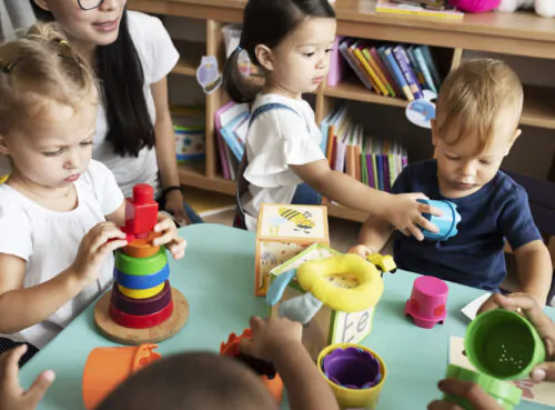 Des enfants s'amusent autour d'une table avec des jouets sous le regard d'une adulte. On voit derrière une bibliothèque avec des livres.