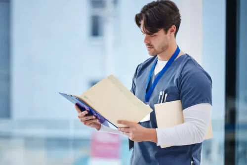 Un infirmier en blouse bleue par-dessus un tee-shirt blanc à manches longues lit un dossier médical ouvert entre ses mains dans un décor d'hôpital.