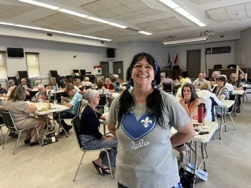 Une femme avec un tee-shirt gris sur lequel est imprimé un coeur bleu avec la fleur de lys blanche du Québec sourit dans une salle où l'on voit de nombreuses personnes assises à des tables, en train de jouer aux cartes.