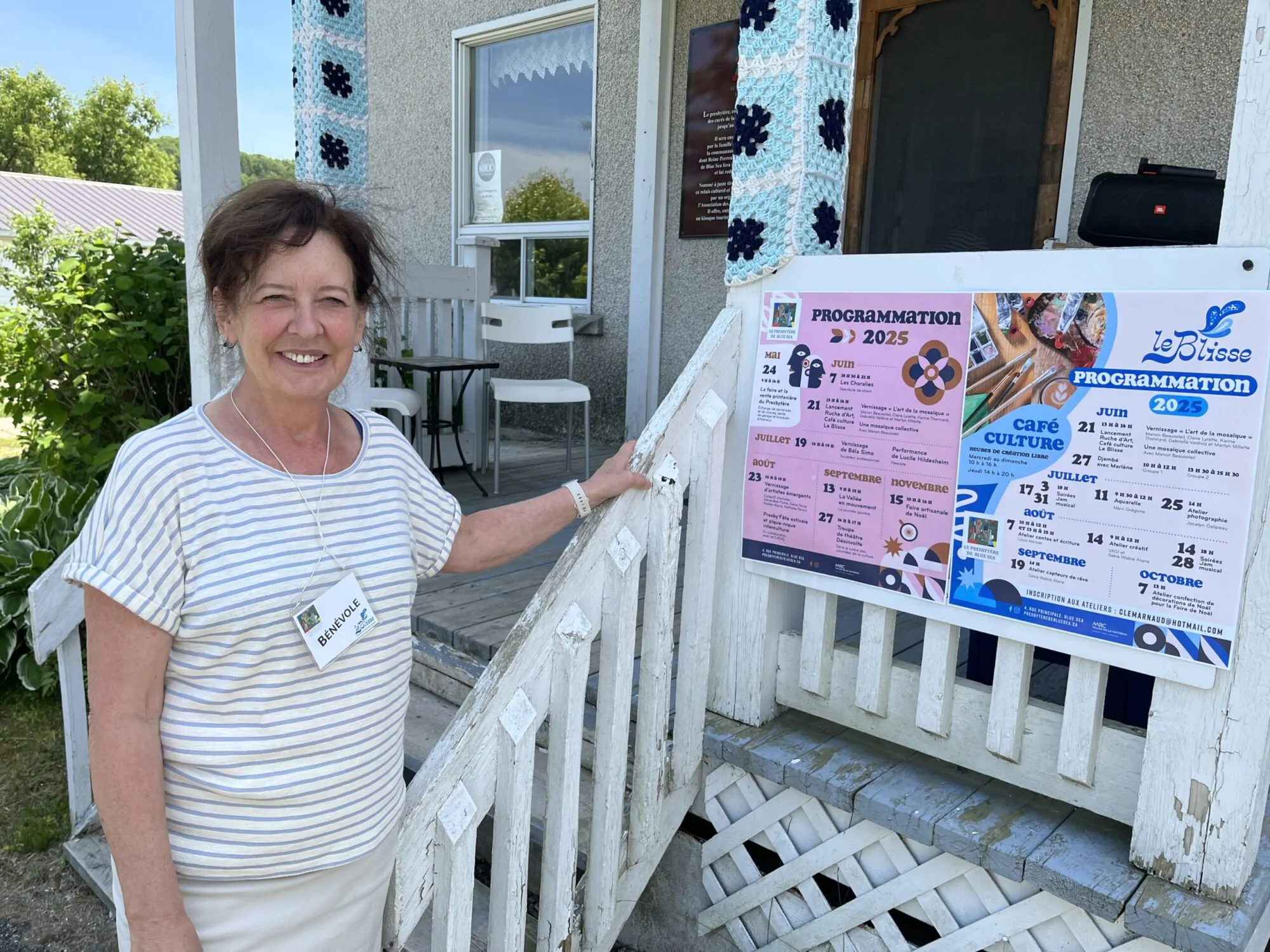 Une femme en habits d'été blancs se tient près de la rampe d'accès et des escaliers d'un bâtiment. Une affiche avec l'annonce d'évènement est collé à la rambarde.