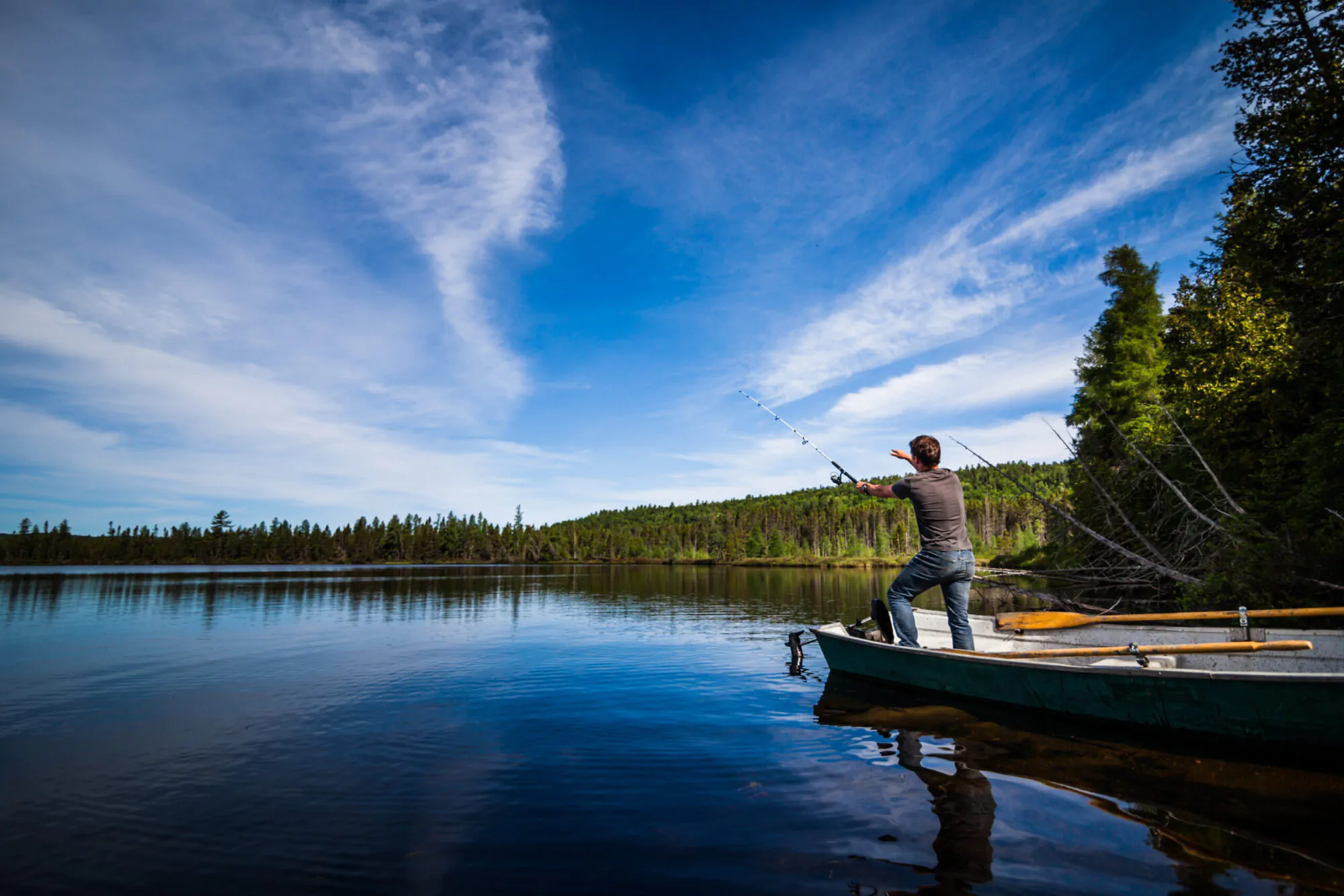 Un homme debout dans sa chaloupe sur un lac calme sous un ciel d'été bleu et légèrement voilé, tient une canne à pêche dans la main et lance sa ligne à l'eau.