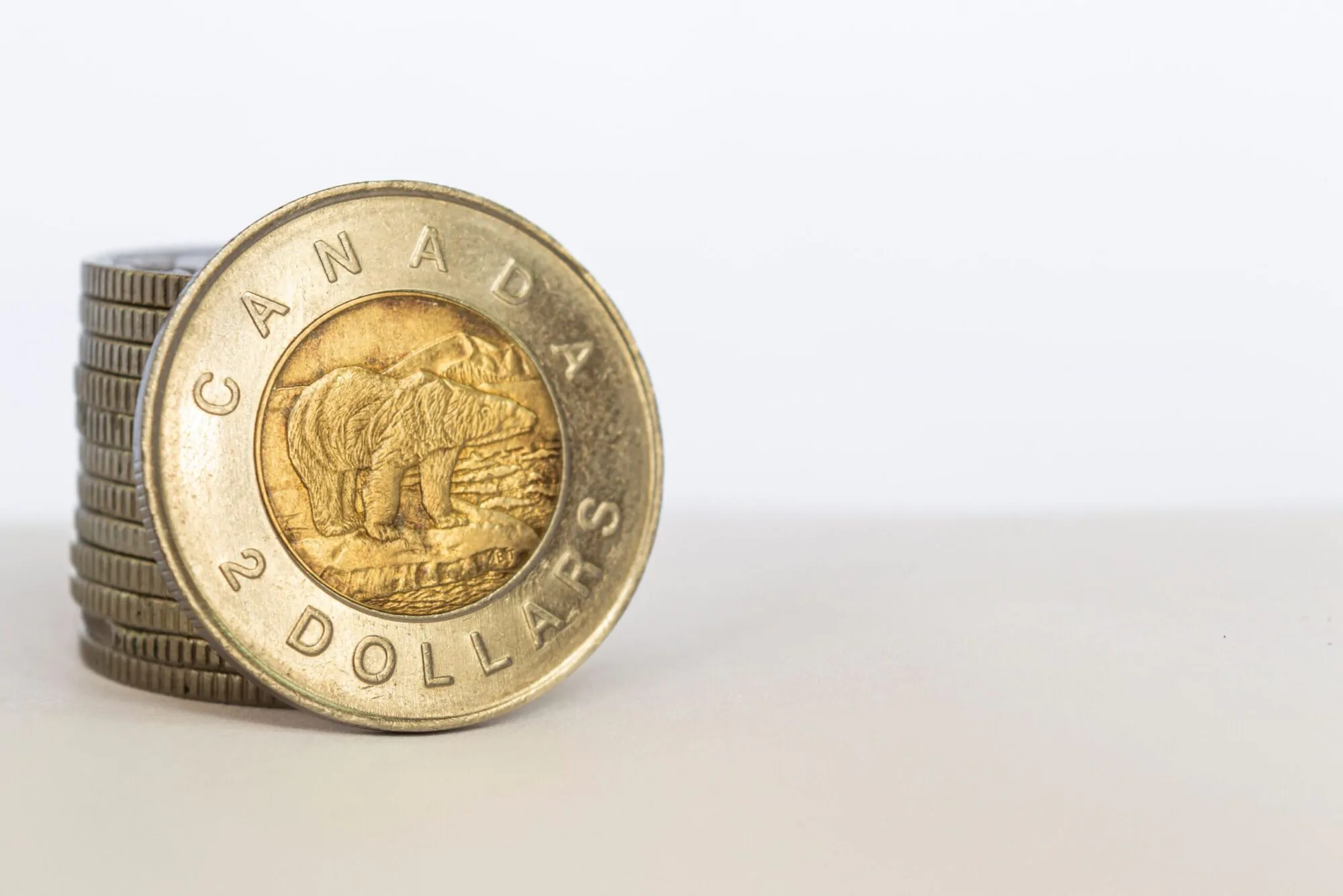 Une pièce de deux dollars canadiens est adossée à une pile de pièces de monnaie sur un fond neutre.