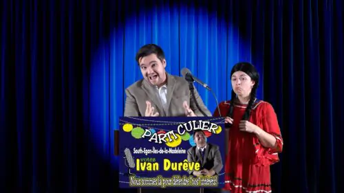 Deux personnes sur une scène, devant un rideau bleu, l’une en costume parlant au micro et l’autre, en robe rouge et tresses. Une affiche humoristique devant le lutrin présente un candidat fictif nommé Ivan Durêve du « Parti Culier », avec le slogan : « Nous sommes là pour défendre nos intérêts ! ».