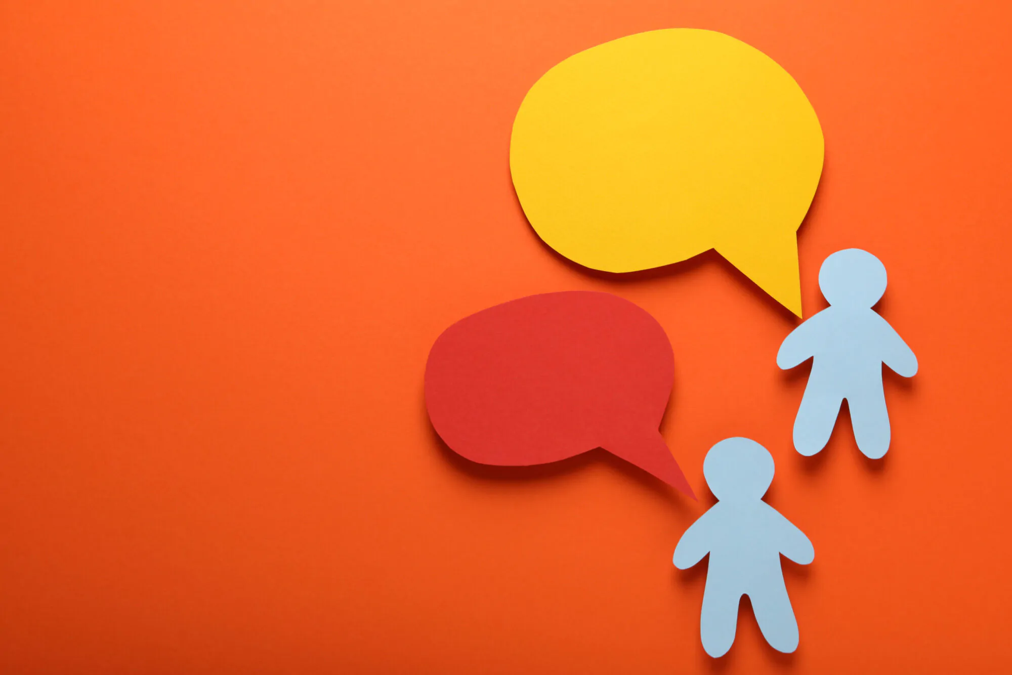 Deux silhouettes humaines stylisées en papier bleu, placées sur un fond orange vif. Chacune est associée à une bulle de dialogue en papier découpé, l'une rouge, l'autre jaune, symbolisant un échange verbal ou une conversation.