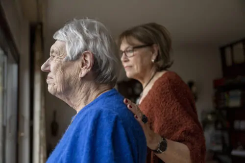 Deux femmes âgées regardent par la fenêtre. Une des aînée a la main posée sur le dos de l'autre, dans un geste affectueux.