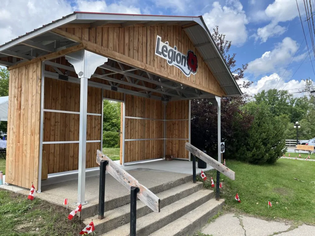 Un abri en bois posé sur une dalle de béton avec des drapeaux canadiens le long des marches et des rampes d'accès, marqué du logo de la Légion royale canadienne et de son coquelicot.