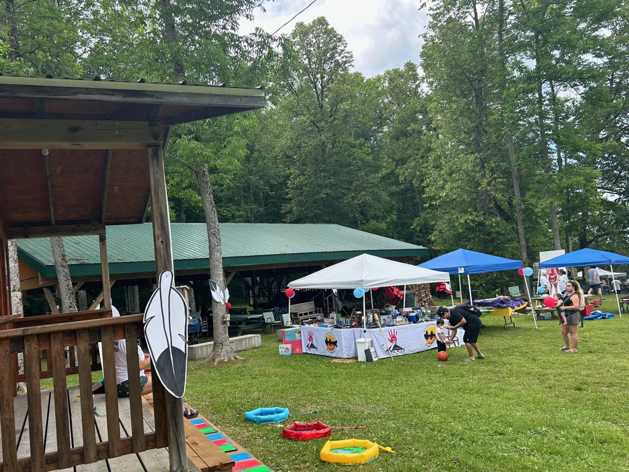 Une fête communautaire en plein air dans un espace boisé, avec des tentes, des tables d’exposition et des décorations festives, comme une plume symbole des Premières Nations. Des familles participent à des activités sur la pelouse.