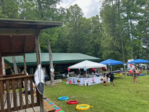 Une fête communautaire en plein air dans un espace boisé, avec des tentes, des tables d’exposition et des décorations festives, comme une plume symbole des Premières Nations. Des familles participent à des activités sur la pelouse.