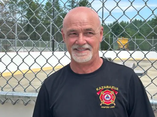 Georges Leblanc, vp Association des pompiers volontaires de Kazabazua, t-shirt noir, logo, barbe blanche et chauve, devant clôture centre communautaire