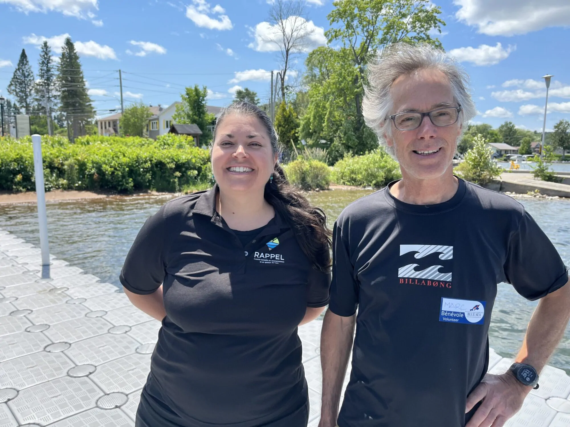 Une femme et un homme se tiennent debout sur un quai public sur un lac du Québec, en bordure du rivage. Ils portent des tee-shirts noirs et ont les cheveux au vent.