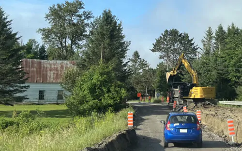 Une voiture bleue s,engage sur une route en reconstruction, bordée de cônes de signalisation orange. Plus loin, des travailleurs et une pelle mécanique jaune s'activent. La route est bordée par un champ, des arbres et une maison. La saison est estivale et le ciel bleu.