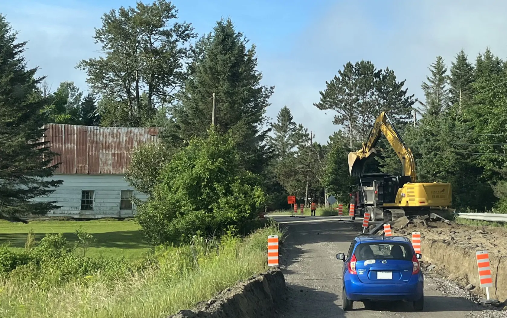 Une voiture bleue s,engage sur une route en reconstruction, bordée de cônes de signalisation orange. Plus loin, des travailleurs et une pelle mécanique jaune s'activent. La route est bordée par un champ, des arbres et une maison. La saison est estivale et le ciel bleu.