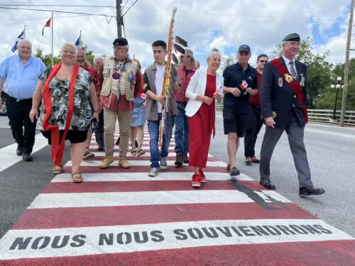 Des hommes et des femmes, certains en tenue civile, d'autres en uniformes, marchent sur une traverse piétonnière rouge et blanche sur laquelle est inscrite la mention " Nous nous souviendrons ".