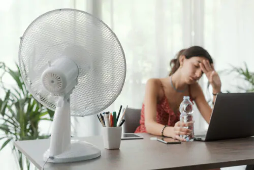 Une femme assise à un bureau devant son ordinateur tient une bouteille d'eau dans une main et son front dans l'autre, tandis qu'un ventilateur en fonction posé sur le bureau envoie de l'air dans sa direction.