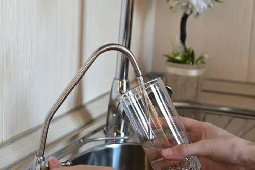 paire de mains remplissant un verre d'eau en vitre à un robinet de cuisine en acier inoxydable