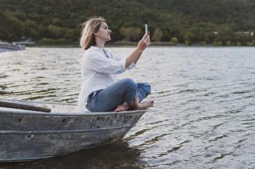 Une jeune femme en chemise blanche et un jean avec un téléphone dans les mains est assise sur un bateau sur la rive de la forêt montagneuse.