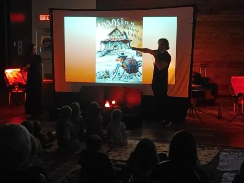 Un homme et une femme présentent un conte pour enfants intitulé "Anansi et la maison hantée", projeté sur un grand écran devant un jeune public attentif. L’ambiance tamisée et les lumières colorées créent une atmosphère immersive et chaleureuse.
