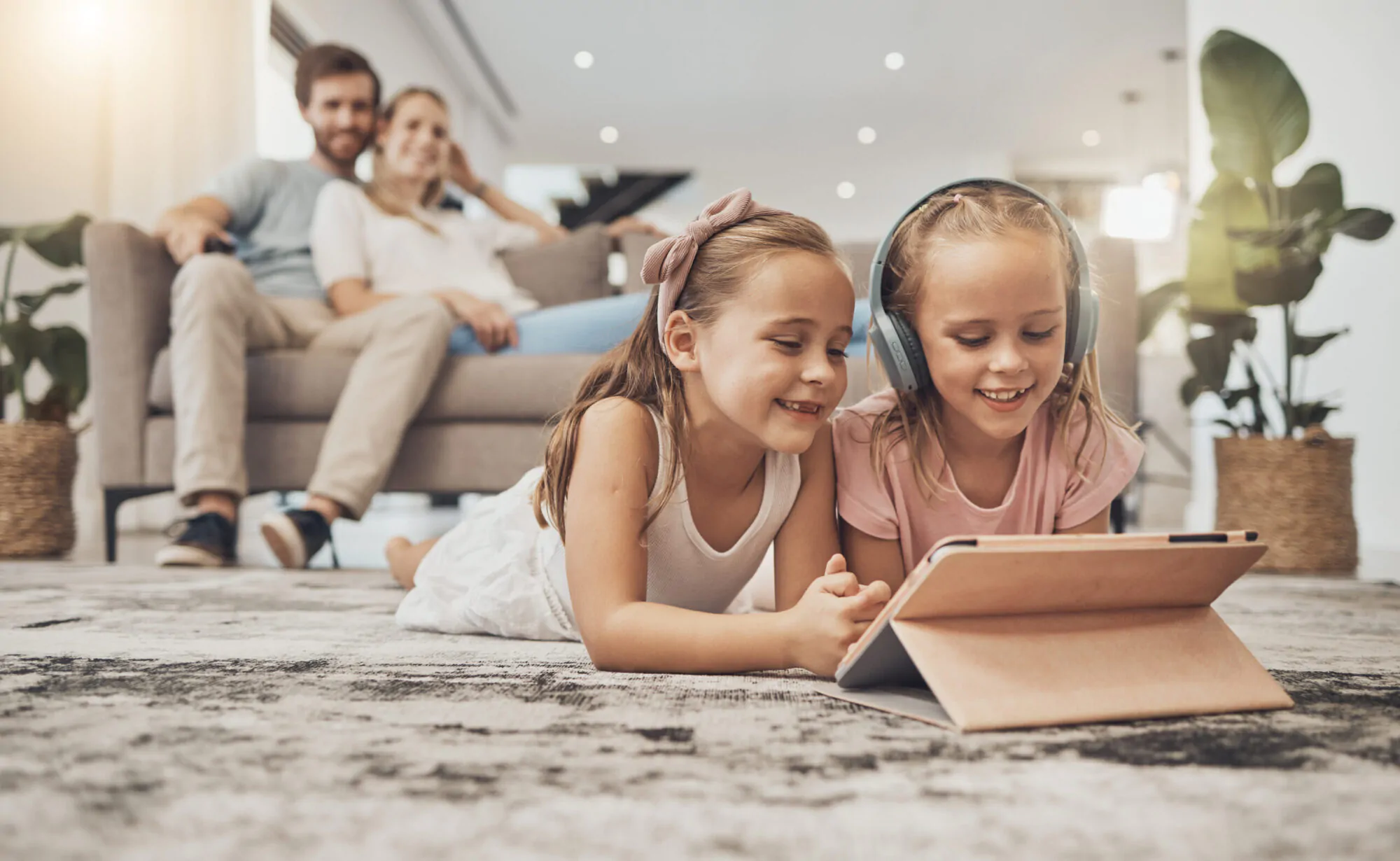 Deux petites filles regardent une tablette allongées dans un salon avec un couple adulte en arrière-plan assis sur un canapé.
