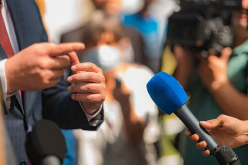 La main d'une journaliste tient un micro, tandis qu'on voit les mains, le veston bleu et la cravate rouge d'un homme politique en train de répondre aux question. On distingue une foule en arrière-plan.