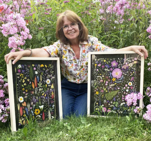 L'artiste Mary Ann McGee souriante à genou sur l'herbe, tenant à sa gauche et à sa droite deux de ses tableaux de fleur séchées. En arrière plan, des fleurs hautes aux pétales roses.