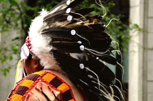 Premières Nations portant une coiffe traditionnelle ornée de plumes, ainsi qu'un vêtement richement décoré de perles et de motifs colorés.