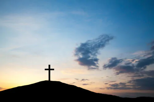 La silhouette d'une croix dressée au sommet d’une colline sur un ciel de coucher de soleil avec des nuages. Les teintes douces de bleu et d’orange créent une atmosphère paisible et spirituelle.