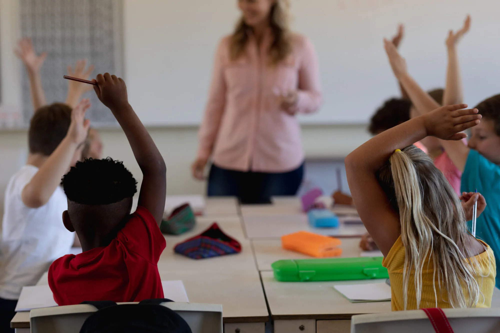 Des écoliers du primaire lèvent la main en classe devant une professeure.