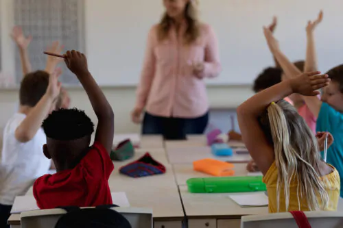 Des écoliers du primaire lèvent la main en classe devant une professeure.