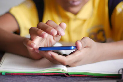 Un élève tient dans sa main un téléphone cellulaire alors qu'un cahier est ouvert devant lui.
