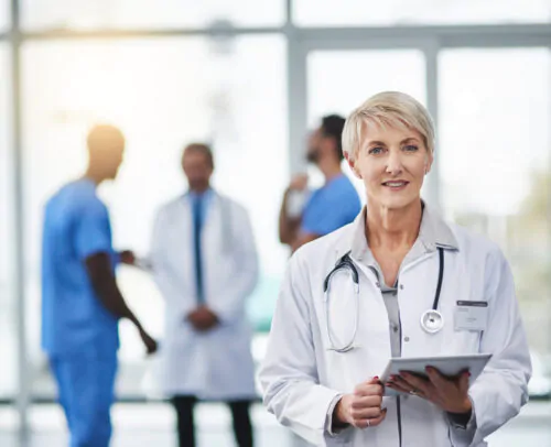 Une médecin âgée se tient debout dans un environnement hospitalier avec du personnel médical en arrière-plan.